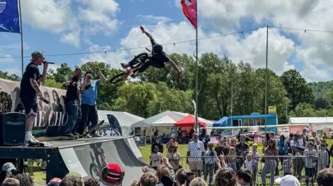 A BMX rider performing an aerial trick over a ramp at an outdoor skate festival.